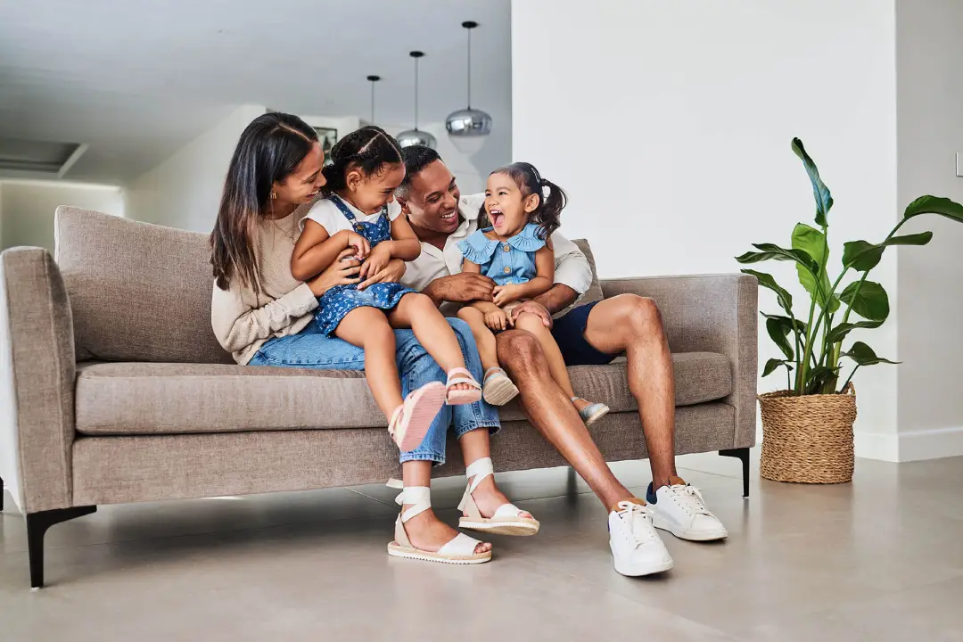 A family of four sitting together on a couch in a cozy living room, surrounded by warm lighting and decorative elements.